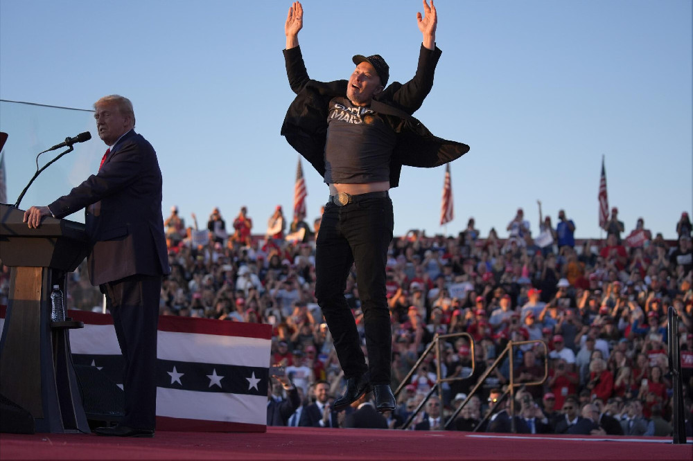 Surrounded by a crowd, Elon Musk leaps into the air behind Donald Trump. 